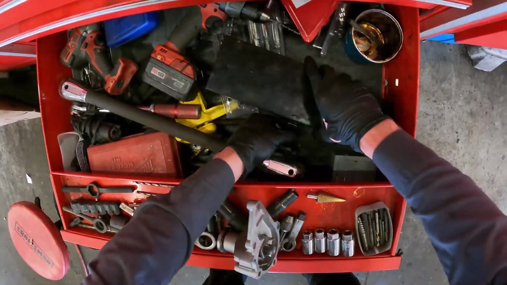 The mechanic removed the trial-fitted replacement water pump, compared its mounting holes with the old pump using drill 