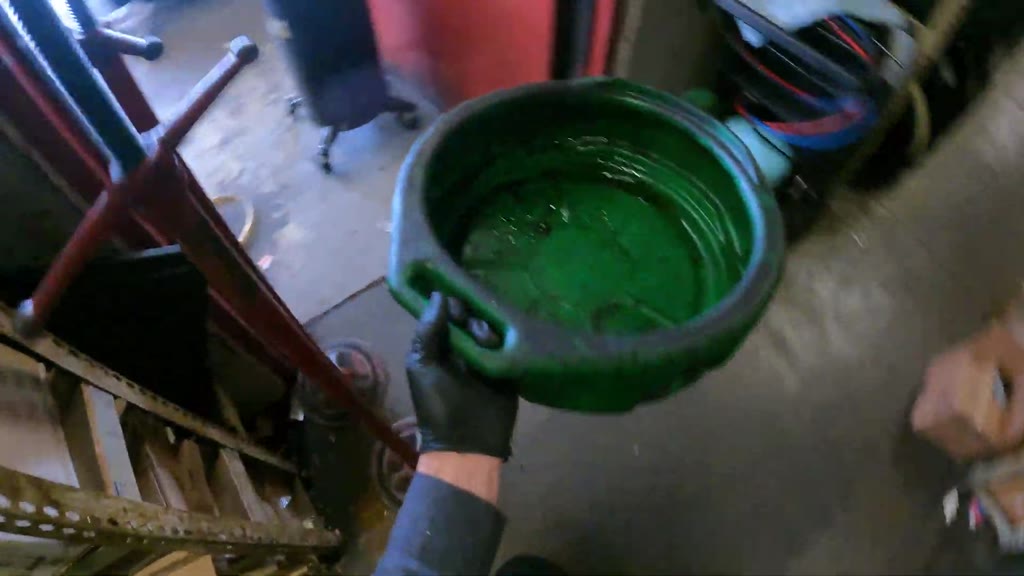 The mechanic retrieved a green drain pan from storage and carried it through the shop to the car’s front bumper.