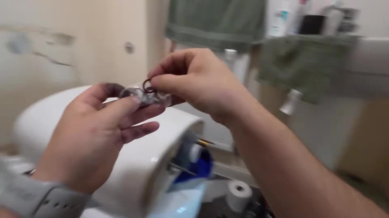 Raw head camera frame: two hands opening a bag of new toilet parts over the tank